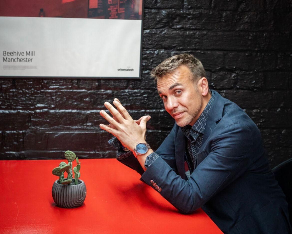 Man sitting at red table gesturing expressively, with small cactus plant, against black brick wall with Beehive Mill Manchester poster.