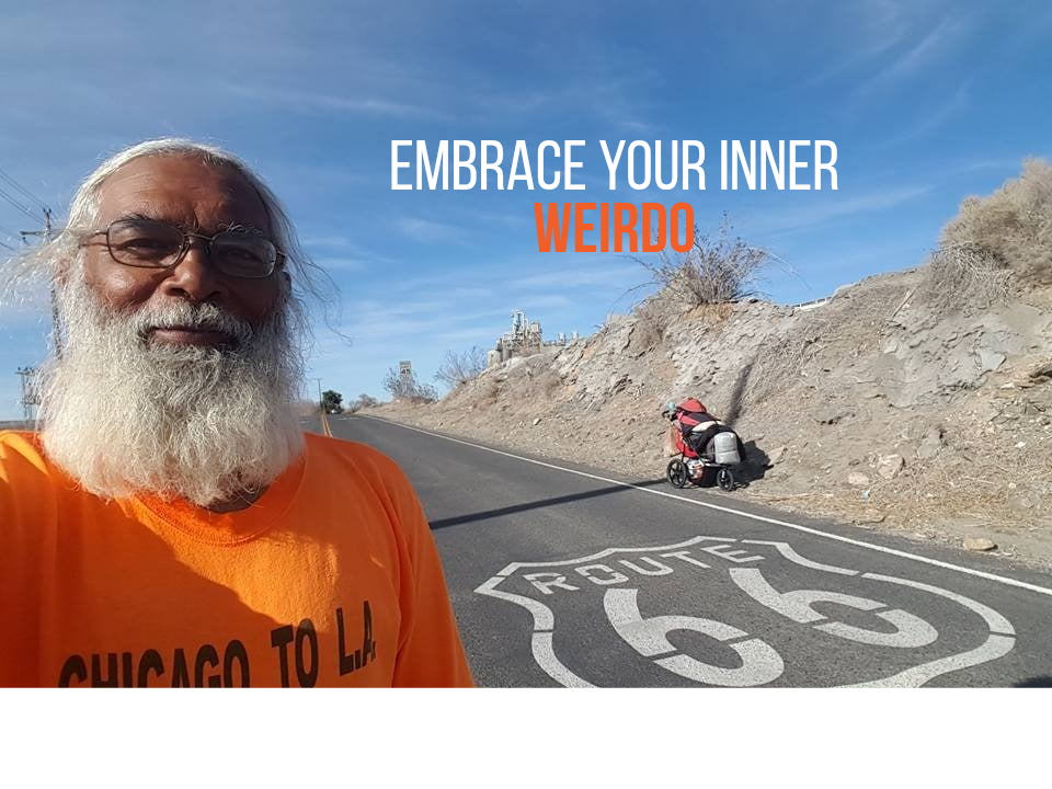 Elderly man in orange shirt standing on Route 66 with motorcycle in background under desert landscape and blue sky