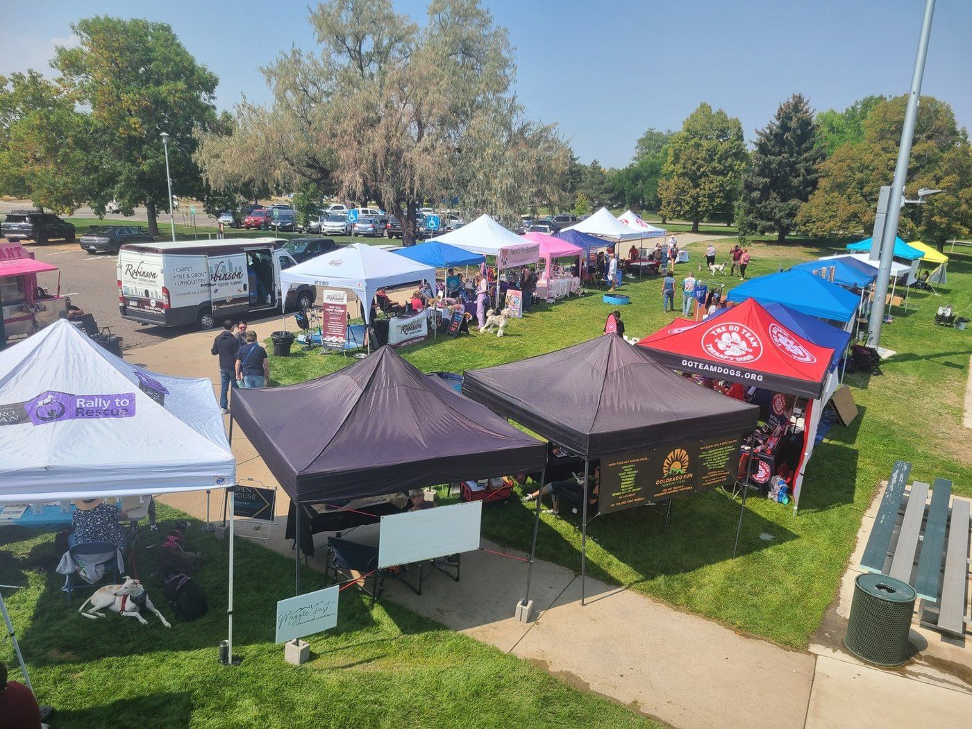 Vendor tents, a work van and dogs on a park lawn at a community dog event, seen from above.