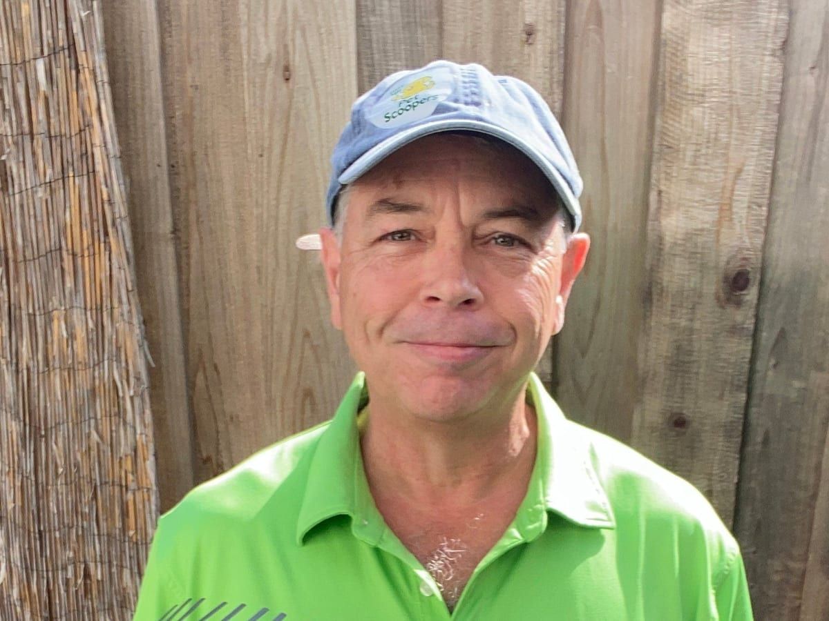 The owner in a lime green Pet Scoopers polo and a blue cap, standing in front of a wooden fence.