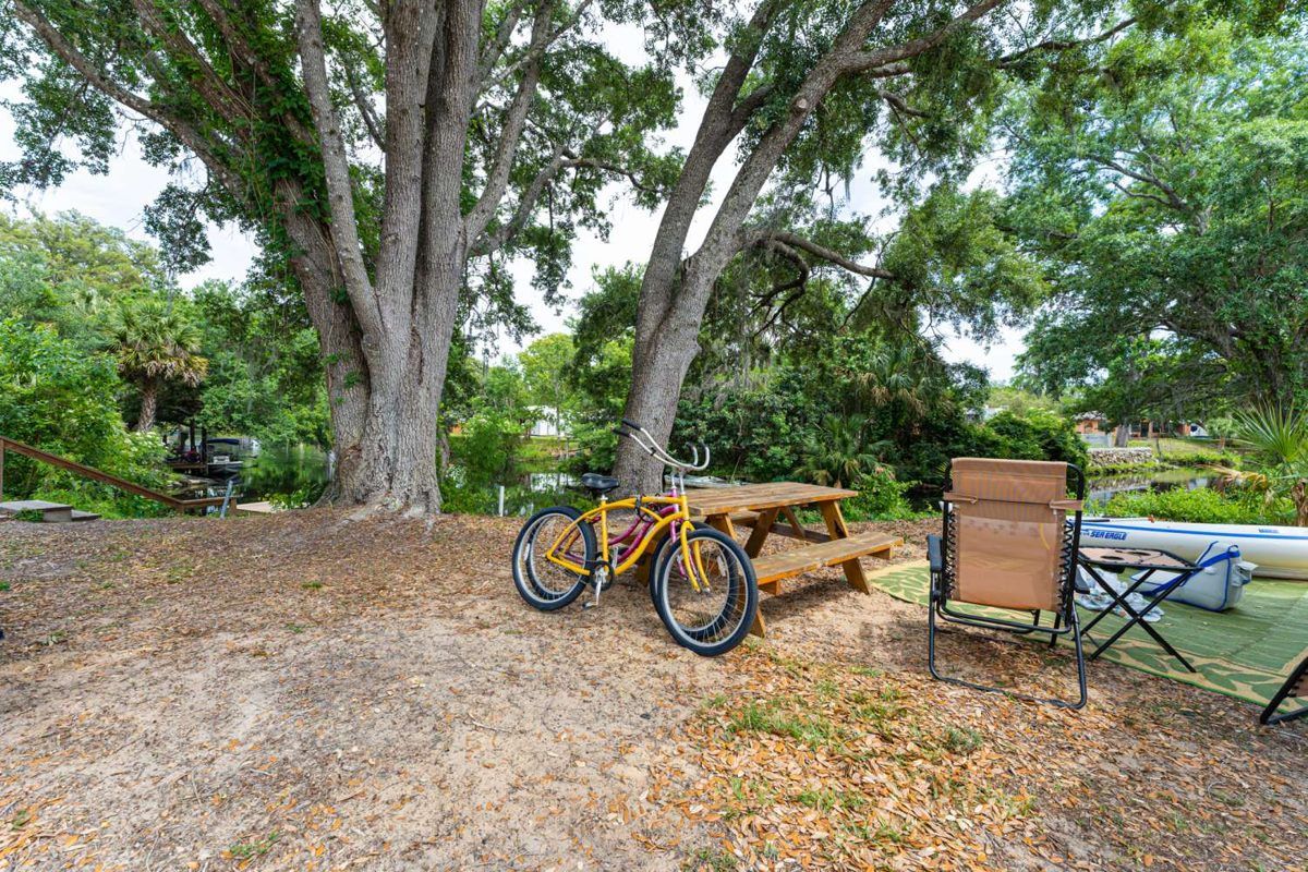 A waterfront RV site with bikes, chairs and a picnic table under oaks