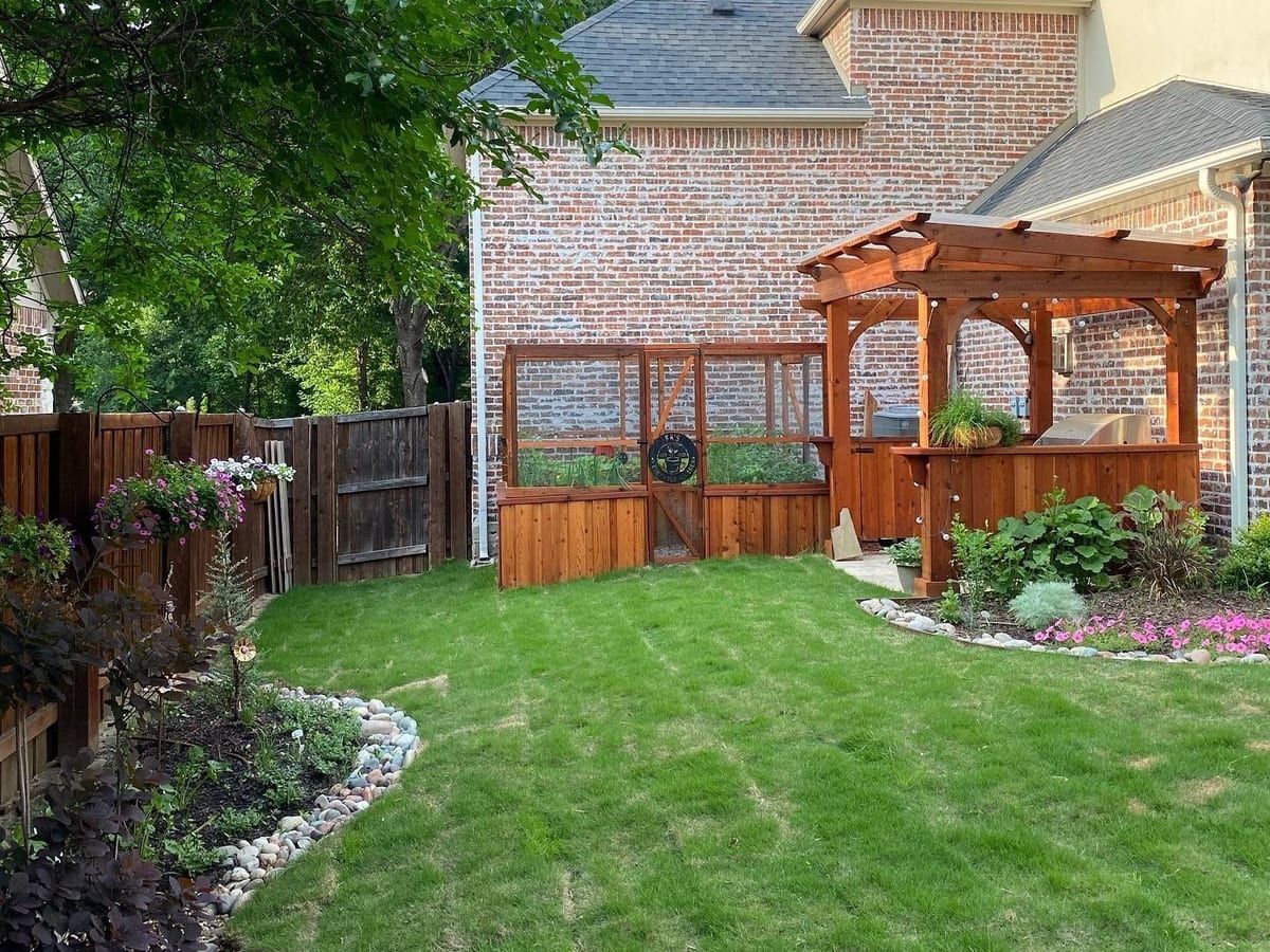 A backyard with a wooden pergola, a flower bed and a trimmed lawn beside a brick house.