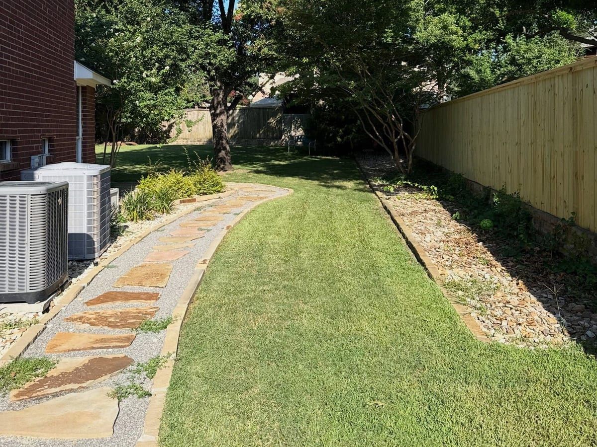 A side yard with a flagstone path, a green lawn, air conditioner units and a wood fence.