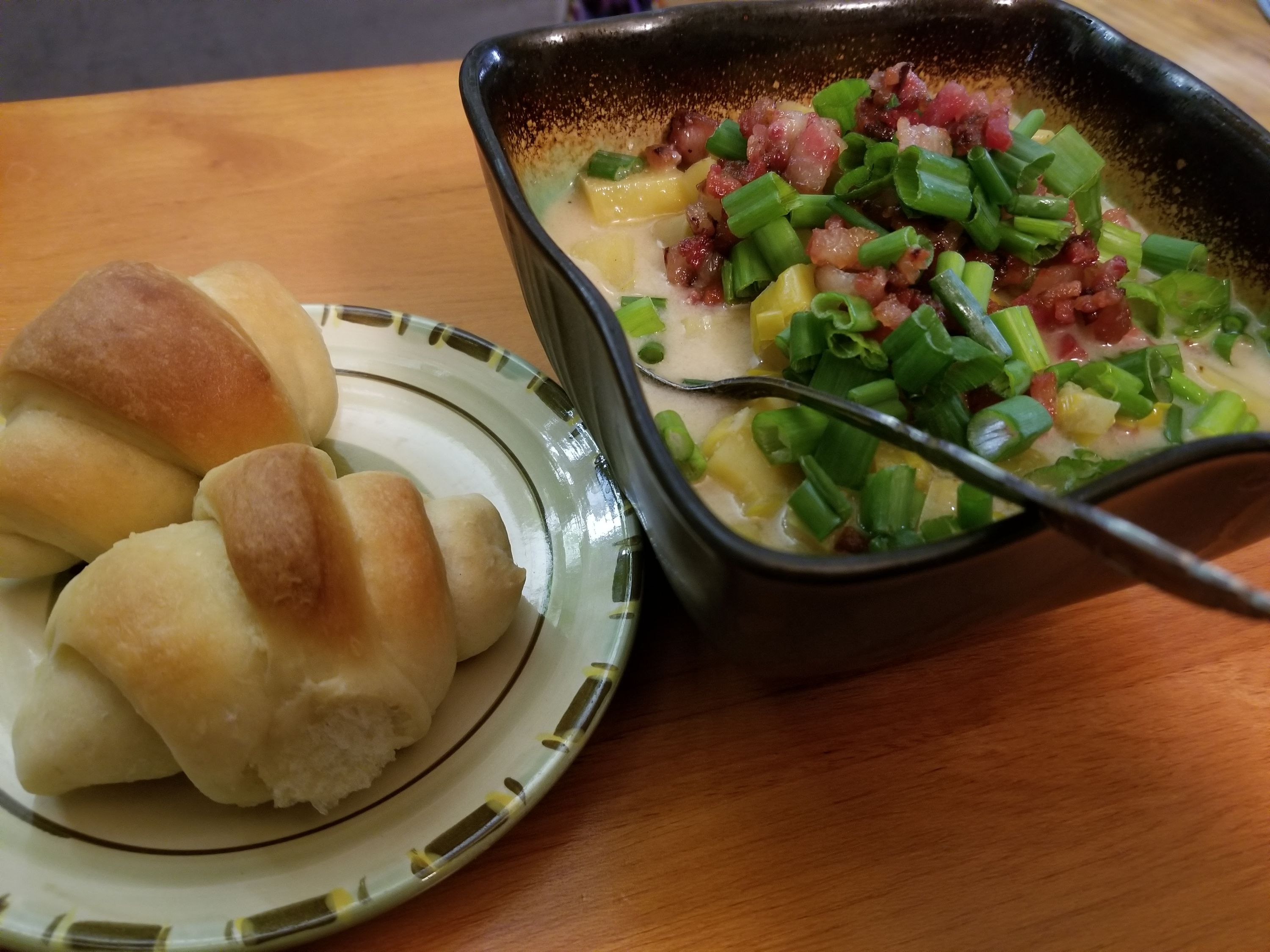 Bowl of creamy soup with diced bacon and green onions, served with sliced bread rolls on a plate beside it